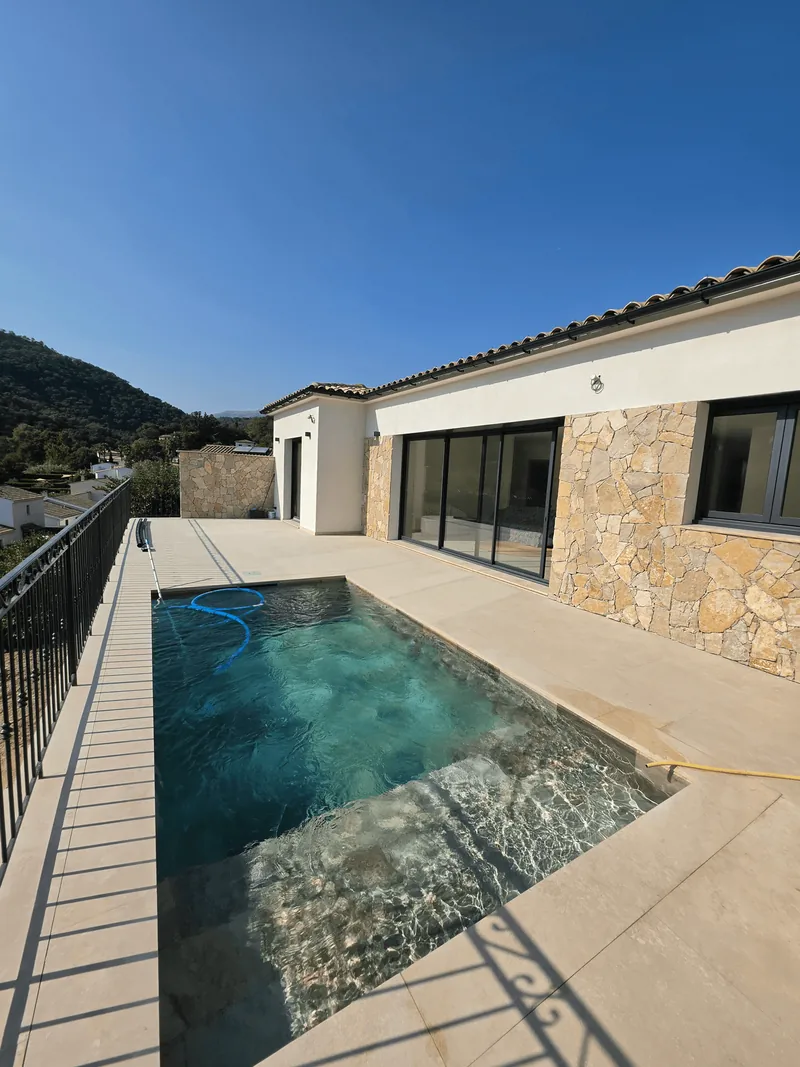 Piscine rectangulaire et terrasse au revêtement clair devant une maison.